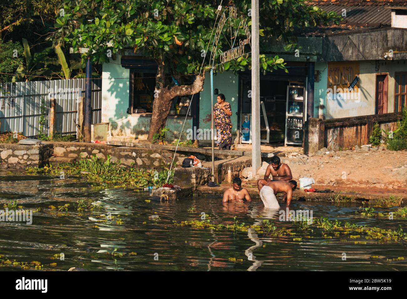 Tre uomini bagnano fuori casa nelle acque posteriori del Kerala, India, in una serata di sole Foto Stock