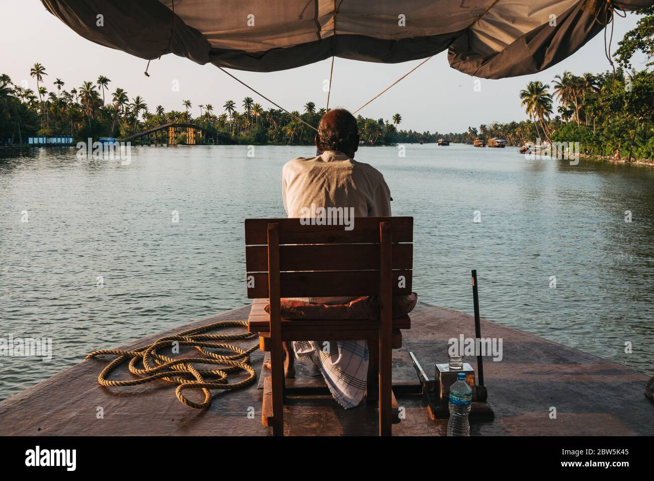 L'autista di una casa galleggiante Keralan guida la barca da un sedile sulla prua, come il sole tramonta Foto Stock