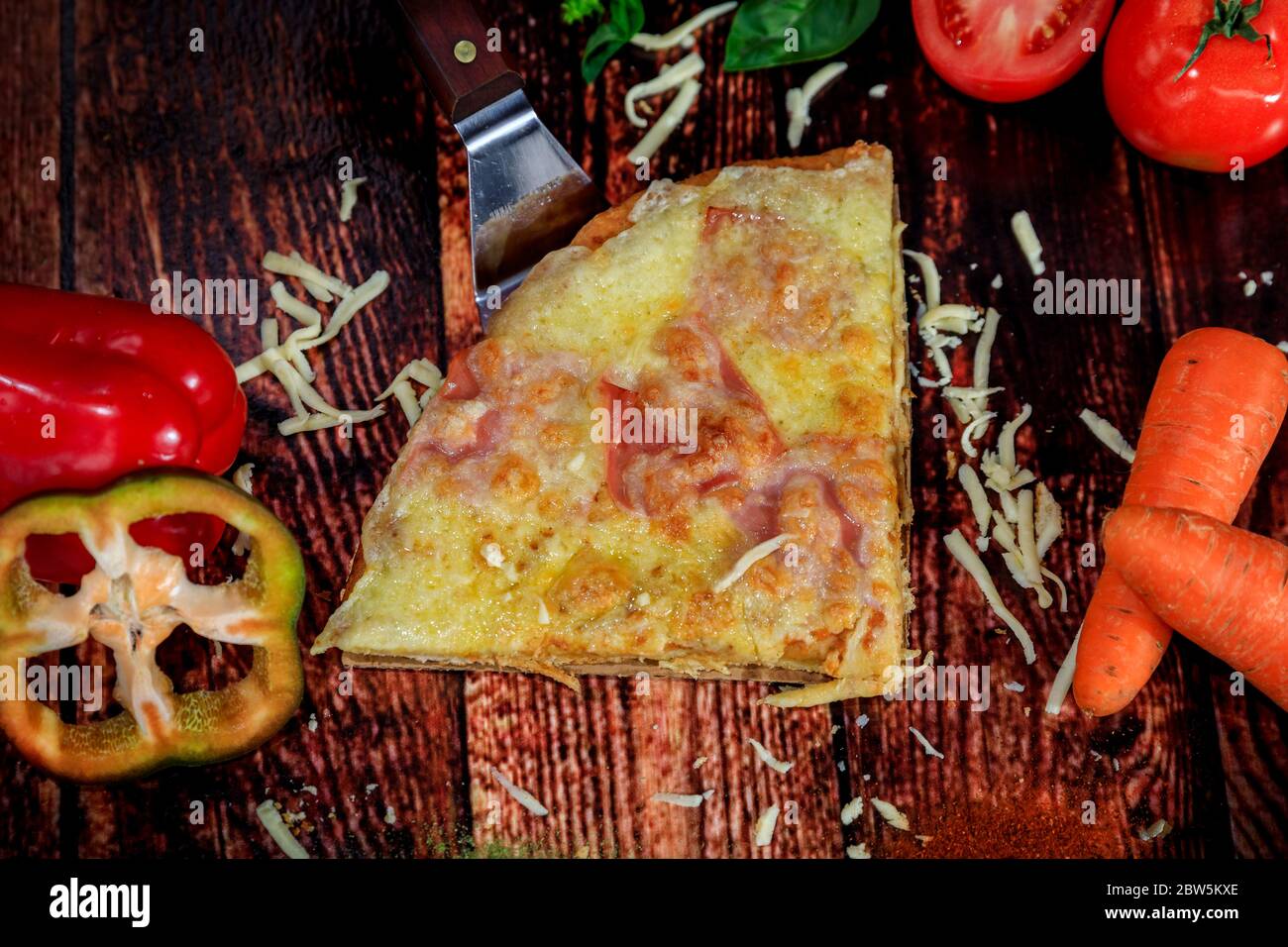 Vista dall'alto della fetta di pizza con formaggio, prosciutto e salame su sfondo tradizionale di legno scuro circondato da ingredienti freschi e verdure Foto Stock