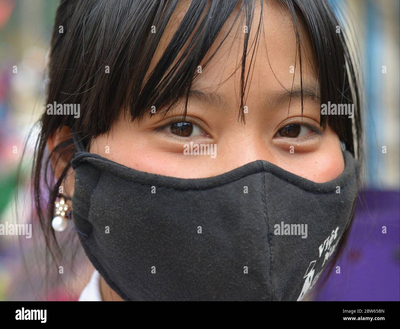 Ragazza asiatica con gli occhi belli copre il naso e la bocca con una maschera riutilizzabile, lavabile in cotone nero. Foto Stock