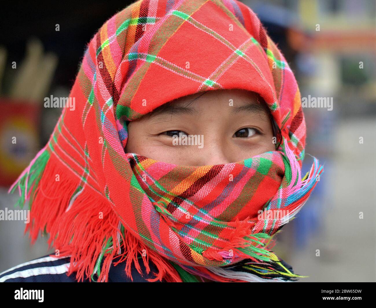 La ragazza hungong vietnamita con occhi asiatici sorridenti ricopre il naso e la bocca con una sciarpa rossa a scacchi. Foto Stock