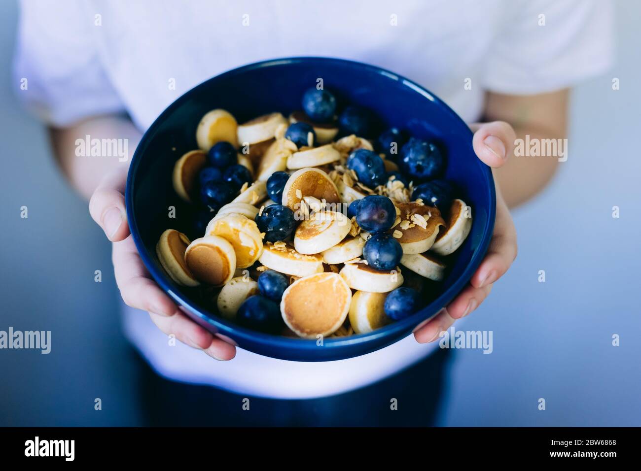 Primo piano di mini-pancake al cereali, mini pancake in una ciotola blu scuro con miele di sirup d'acero e mirtillo nelle mani dei bambini. Cibo di fondo. Foto Stock