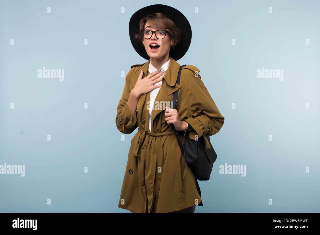 Giovane donna sorpresa con capelli corti scuri in trench cappotto, occhiali  e cappello con zaino stupito guardando da parte su sfondo blu isolato Foto  stock - Alamy