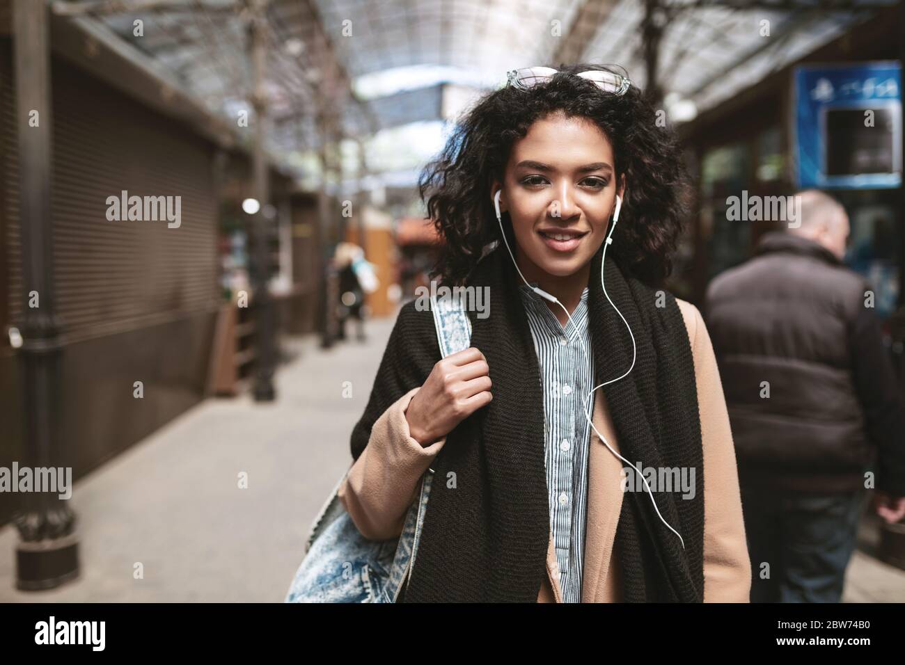 Ritratto di bella ragazza afroamericana in piedi sulla strada e guardando in macchina fotografica Cool ragazza con capelli ricci scuri in auricolari che girano per la città Foto Stock