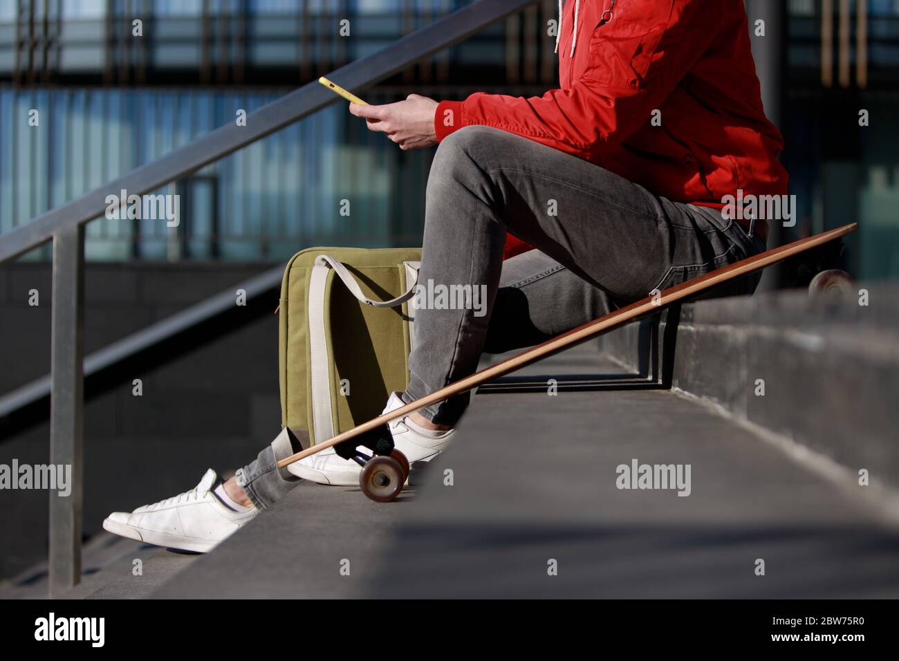 Elegante uomo longboarder in abiti casual con il suo smartphone, appoggiato sui gradini, seduto con longboard / skateboard all'aperto, vista laterale, taglio i Foto Stock