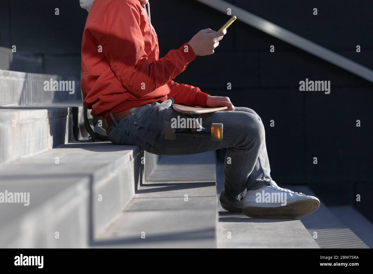 Elegante uomo longboarder in abiti casual con il suo smartphone, appoggiato sui gradini, seduto con longboard / skateboard all'aperto, vista laterale, taglio i Foto Stock