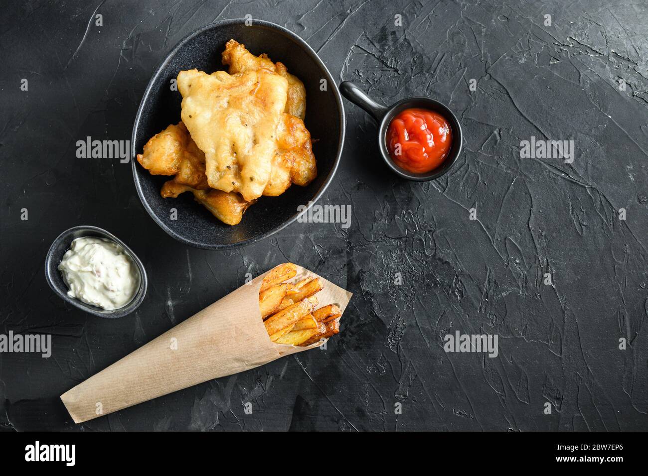 Pesce e patatine in un tuffo di carta cono e limone - merluzzo fritto, patatine fritte, fette di limone, salsa tartaro, ketchup tomatoe piselli funghi su sfondo nero Foto Stock