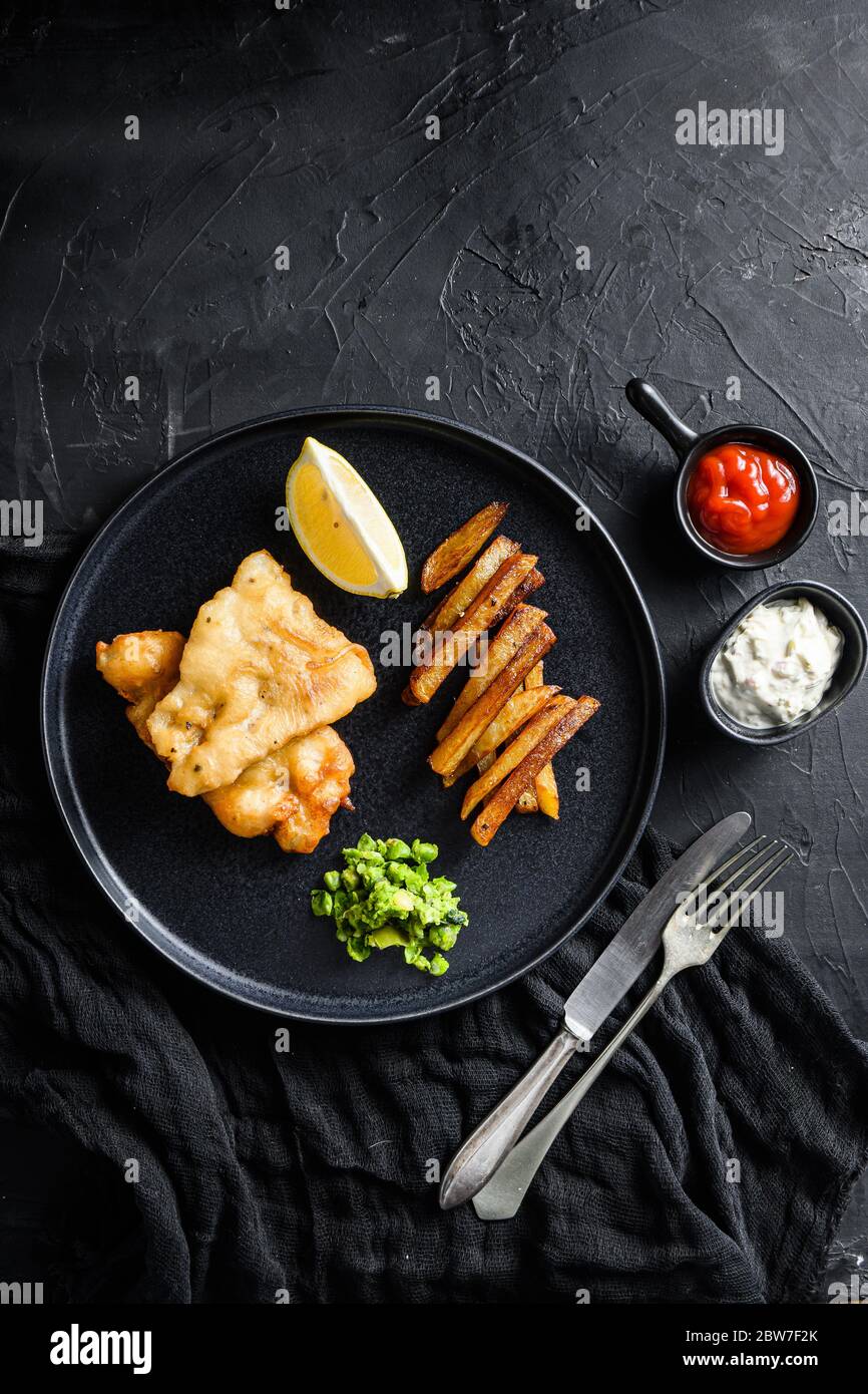 Pesce e patatine. Tradizionale piatto britannico di pesce fritto, patatine, piselli minati e salsa tartare e vista ketchup dall'alto, bac in pietra nera Foto Stock