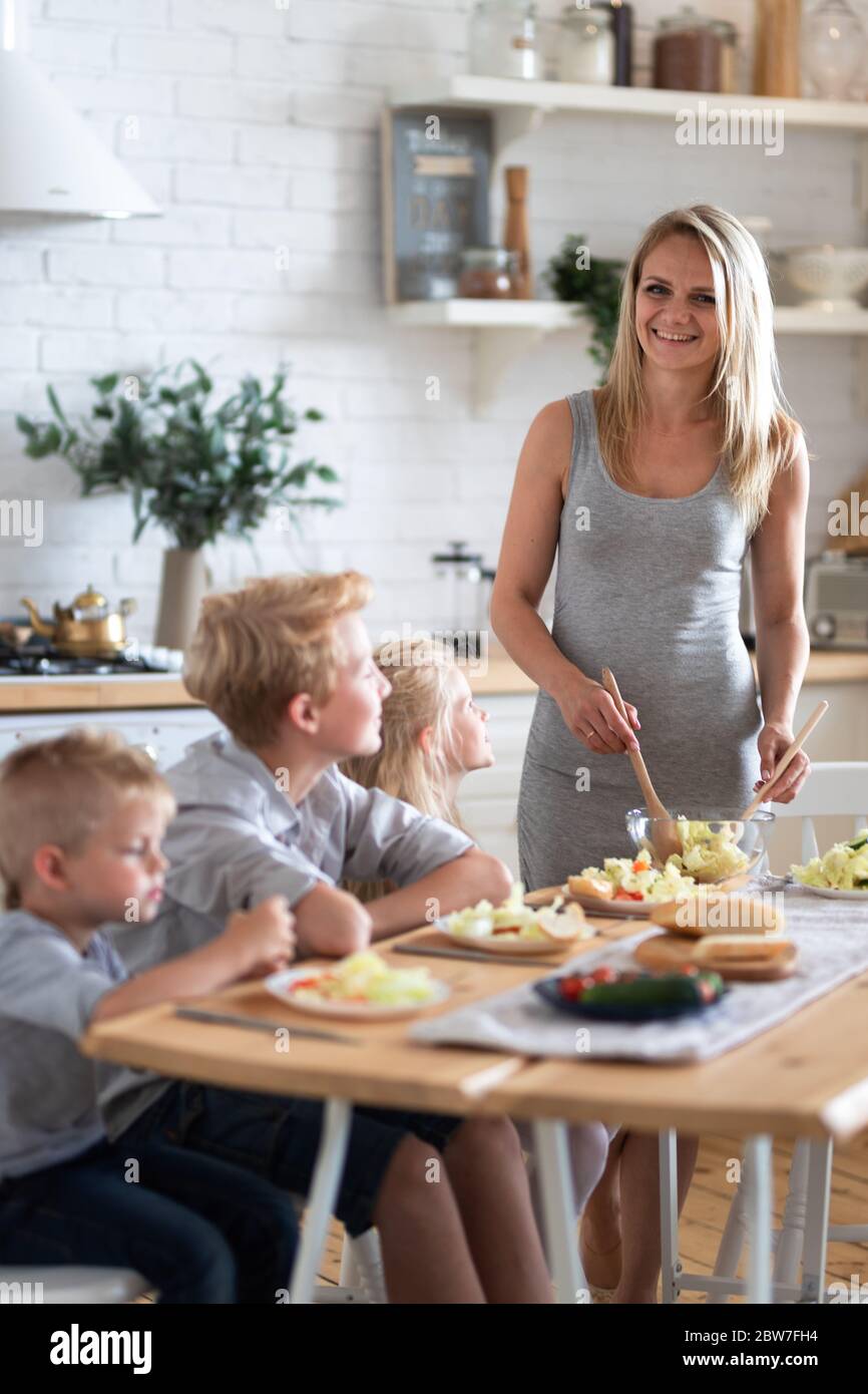 madre bionda di famiglia con tre bambini due figli e figlia che mangiano cibo sano in cucina a casa, mamma mette insalata verde sui piatti per i suoi bambini. Foto Stock