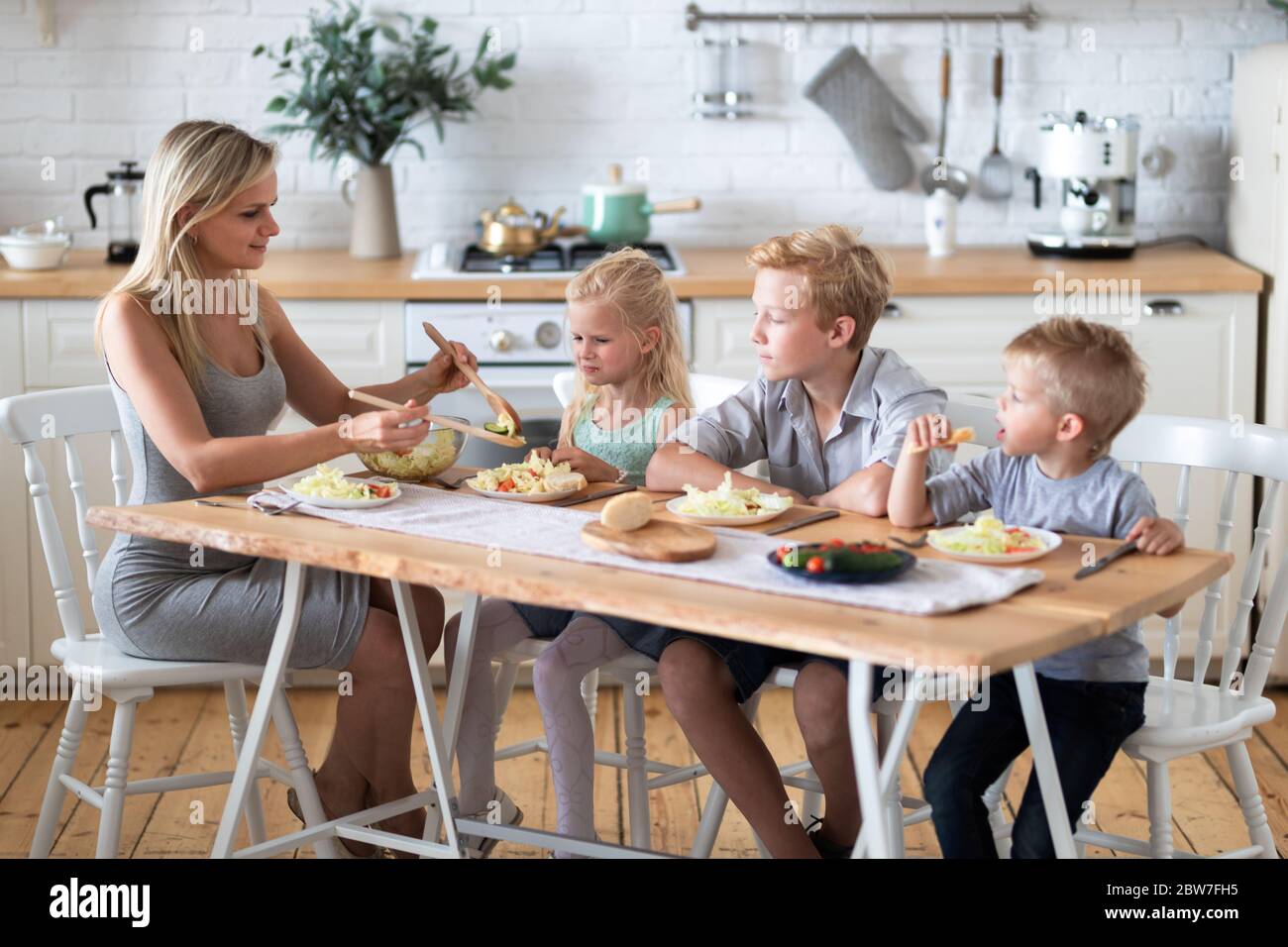 madre bionda di famiglia con tre bambini due figli e figlia che mangiano cibo sano in cucina a casa, mamma mette insalata verde sui piatti per i suoi bambini. Foto Stock