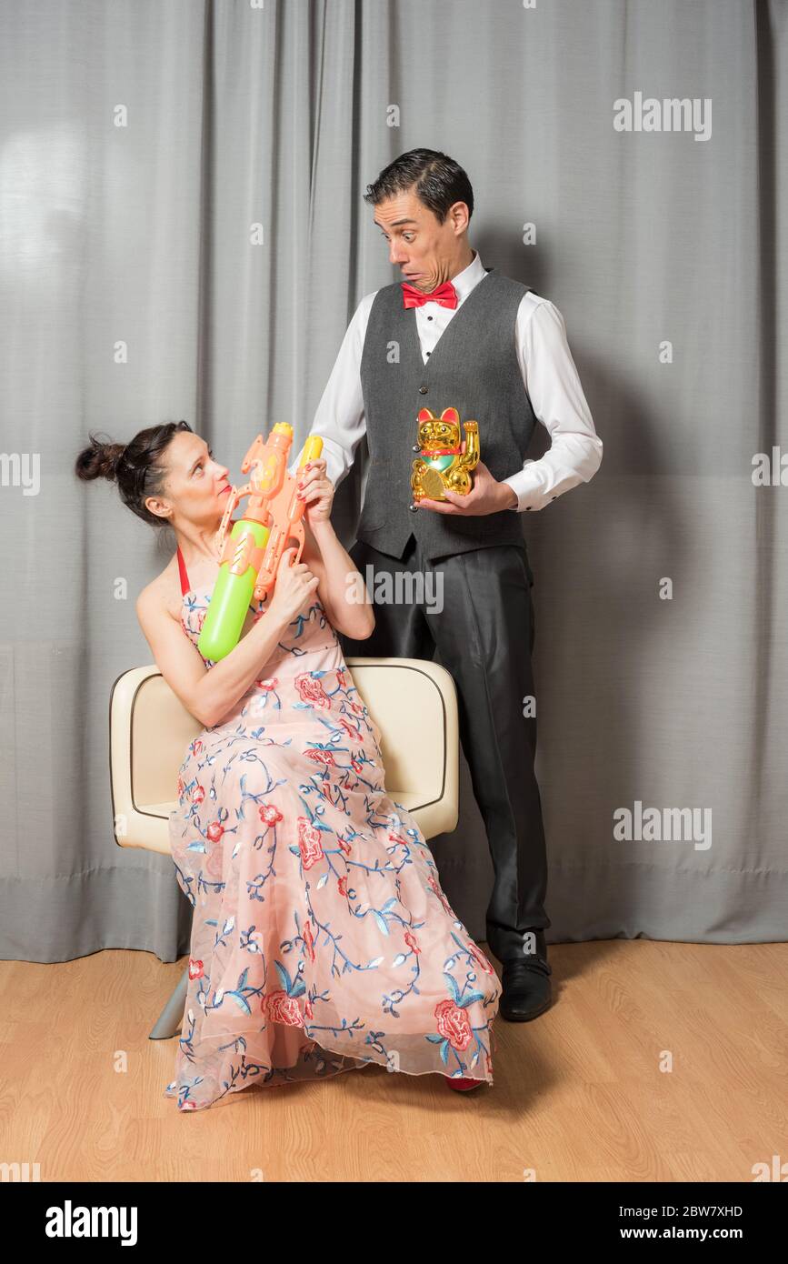 Ritratto di matrimonio sorridente ed eccentrico. La donna, seduta in un  elegante abito fiorito, che porta un'arma giocattolo. L'uomo, in piedi,  indossa un'elega Foto stock - Alamy