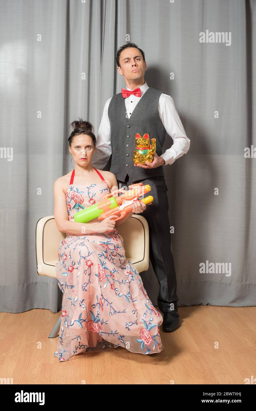 Eccentrici ritratti di matrimonio con atteggiamento solenne. La donna, seduta in un elegante abito fiorito, che porta un'arma giocattolo. L'uomo, in piedi, indossando un Foto Stock