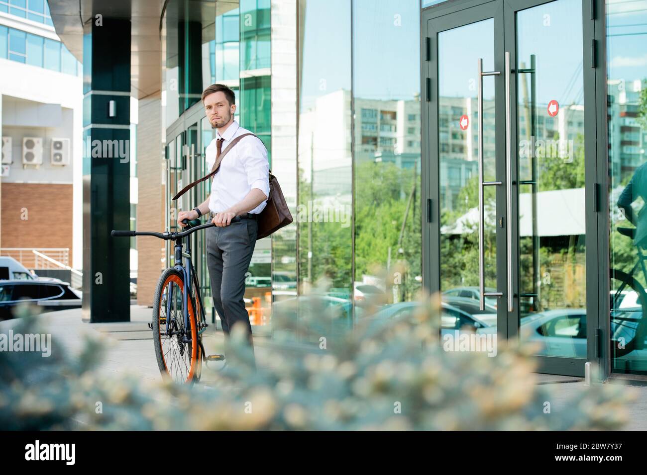 Giovane direttore d'ufficio contemporaneo in abbigliamento da formalwear in piedi accanto al centro affari mentre andando a tornare a casa in bicicletta Foto Stock
