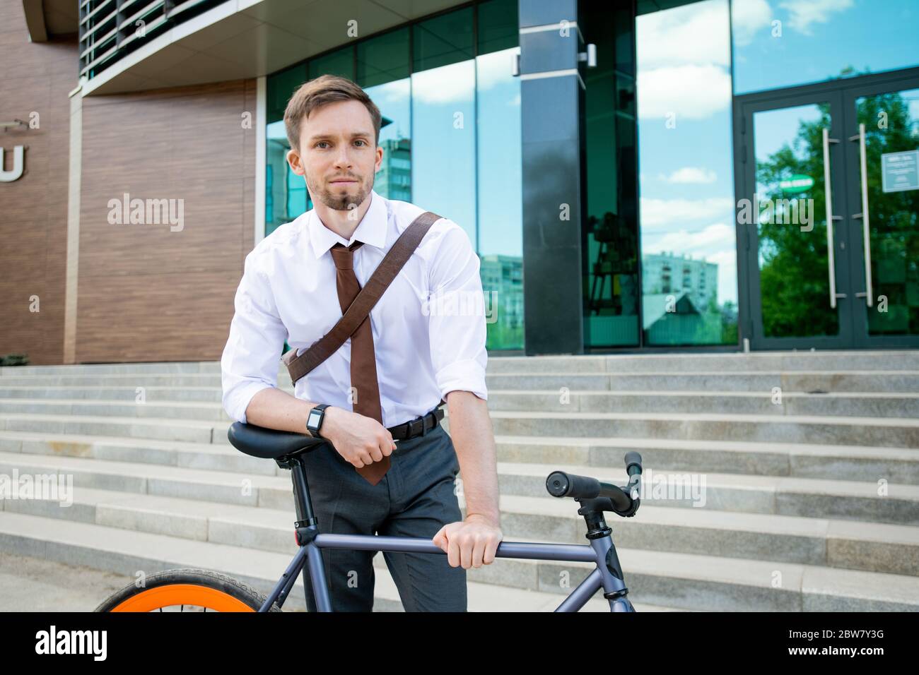 Felice giovane uomo d'affari riuscito che ti guarda mentre si appoggia sulla sua bicicletta sullo sfondo di edificio esterno e scala Foto Stock