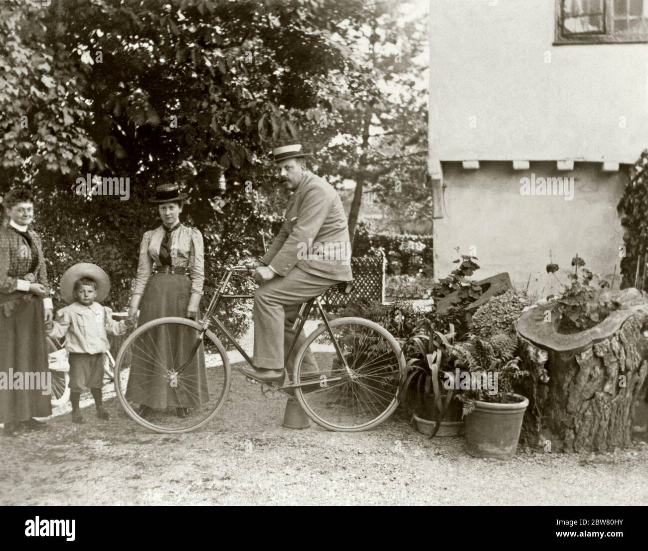 Un uomo si pone sulla sua moto (equilibrandosi con l'aiuto di un vaso di fiori capovolto) mentre altri membri della sua famiglia guardano sopra, Gloucestershire, Inghilterra, Regno Unito c. 1900. L'uomo indossa un vestito e un cappello di paglia, come una donna e un ragazzino. Le persone raffigurate sono lavoratori agricoli. Erano anche appassionati giardinieri con le loro piante in vaso in mostra - notare l'uso di tronchi di albero scavati fuori per fare piantatrici. Foto Stock