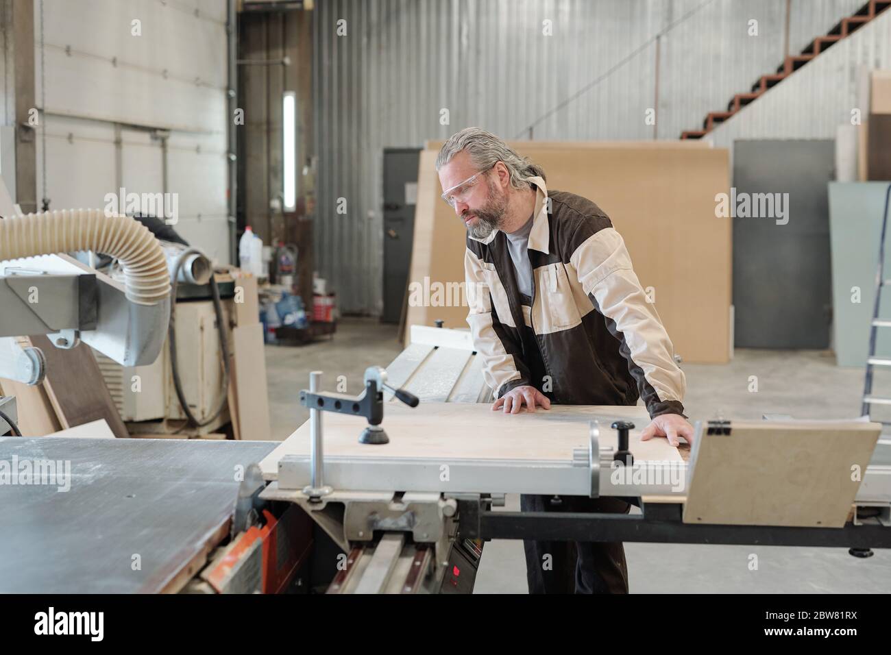 Lavoratore senior di mobili che produce fabbrica di fissaggio di pannelli rettangolari su banco di lavoro prima di tagliarli o smerigliarli con utensile manuale elettrico Foto Stock