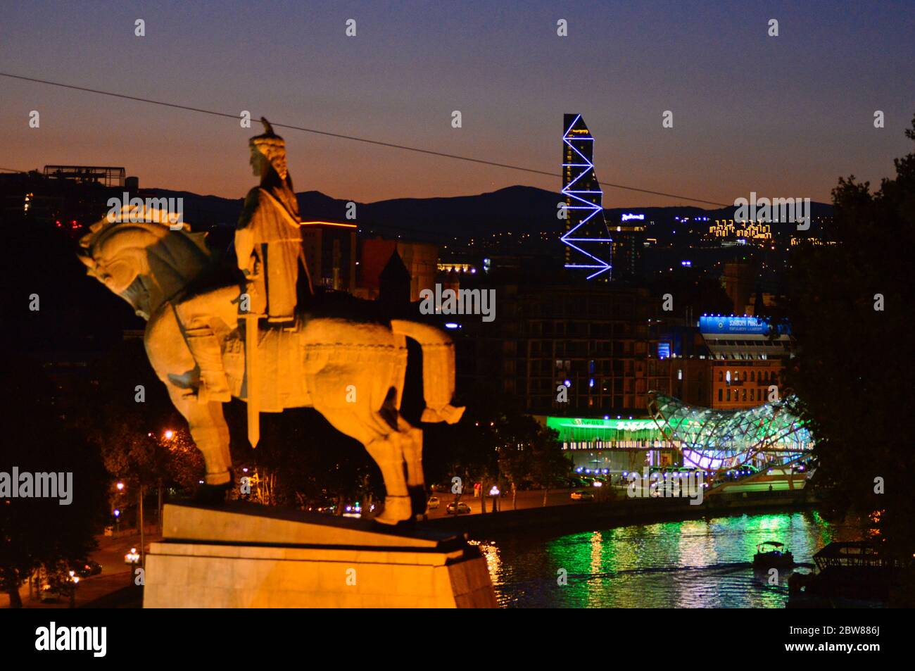 Tbilisi: Monumento del re Vakhtang Gorgasali, al crepuscolo, con il fiume Kura, Ponte della Pace e le montagne sullo sfondo. Georgia Foto Stock