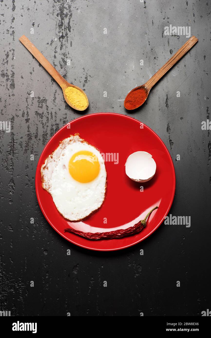 Condire con uova fritte vicino a cucchiai con spezie e peperoncino rosso. Colazione e concetto di arte alimentare. Omelette, gusci e riflessi fanno un'immagine sorridente del volto. Cibo posto su piatto rosso su fondo nero Foto Stock