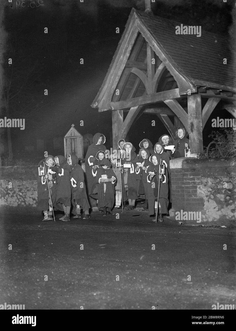 Giovani cantanti carol incappucciati cantano dal cancello del lico fuori della chiesa di Eynsford , Kent . 1935 Foto Stock