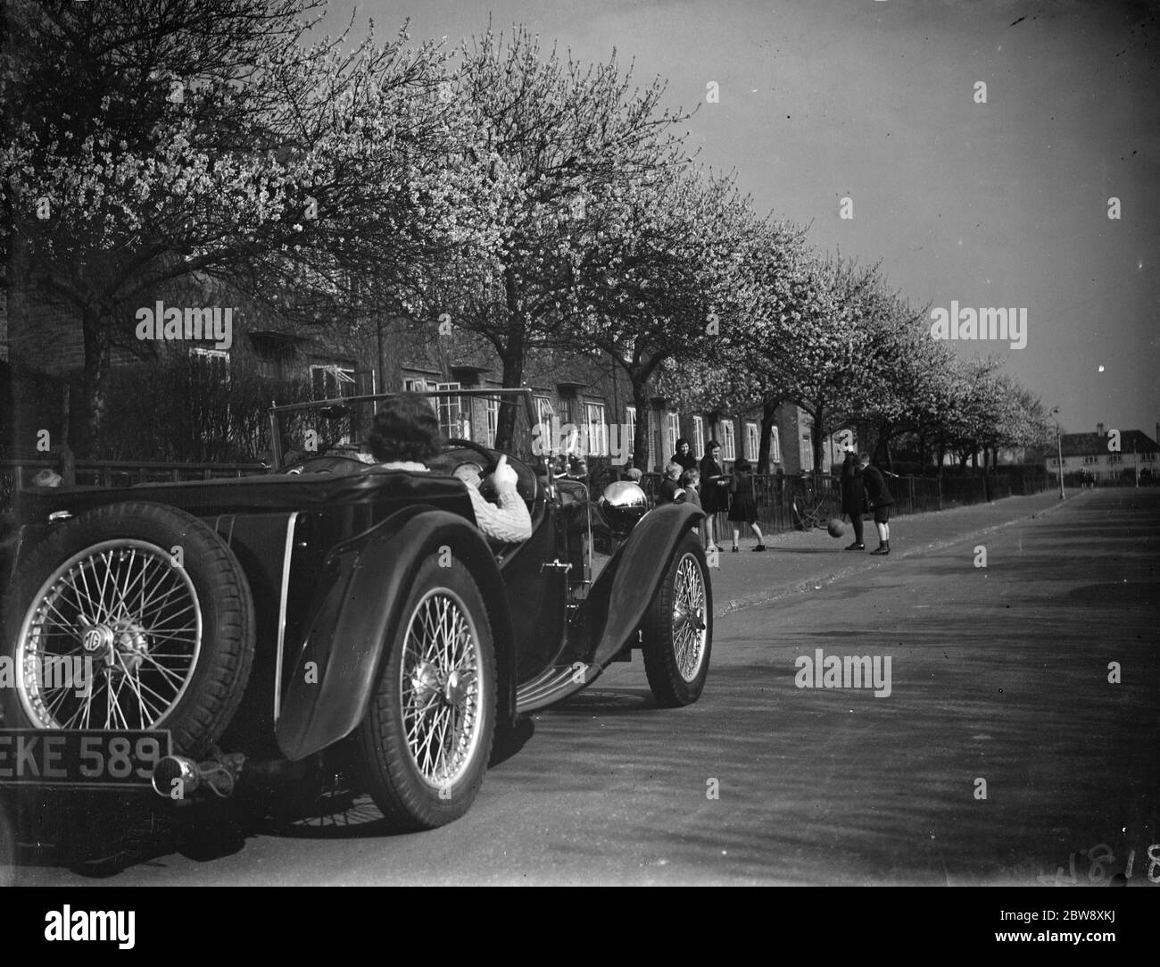 Una macchina sportiva parcheggiata sul lato della strada mentre i bambini giocano sotto gli alberi di mandorle a Crayford . 1938 Foto Stock