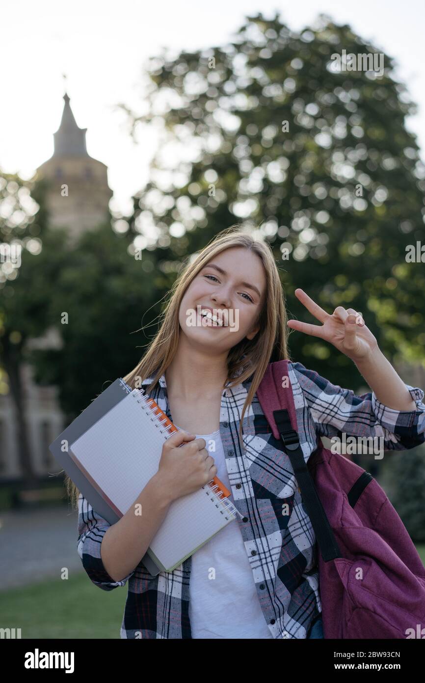 Ragazza allegra che mostra il segno della vittoria. Concetto di ritorno a scuola. Ritratto di studente sorridente con zaino e libri a piedi per l'università Foto Stock