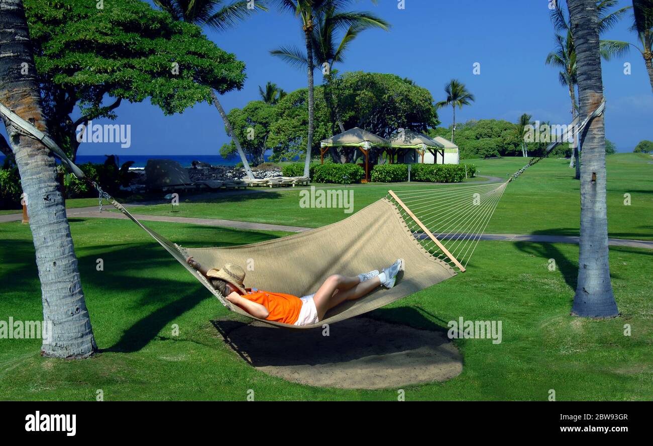 La donna anziana si trova in un'amaca tra due palme in un resort sulla costa di Kohala della Big Island delle Hawaii. Il suo cappello di paglia si stende su di lei Foto Stock