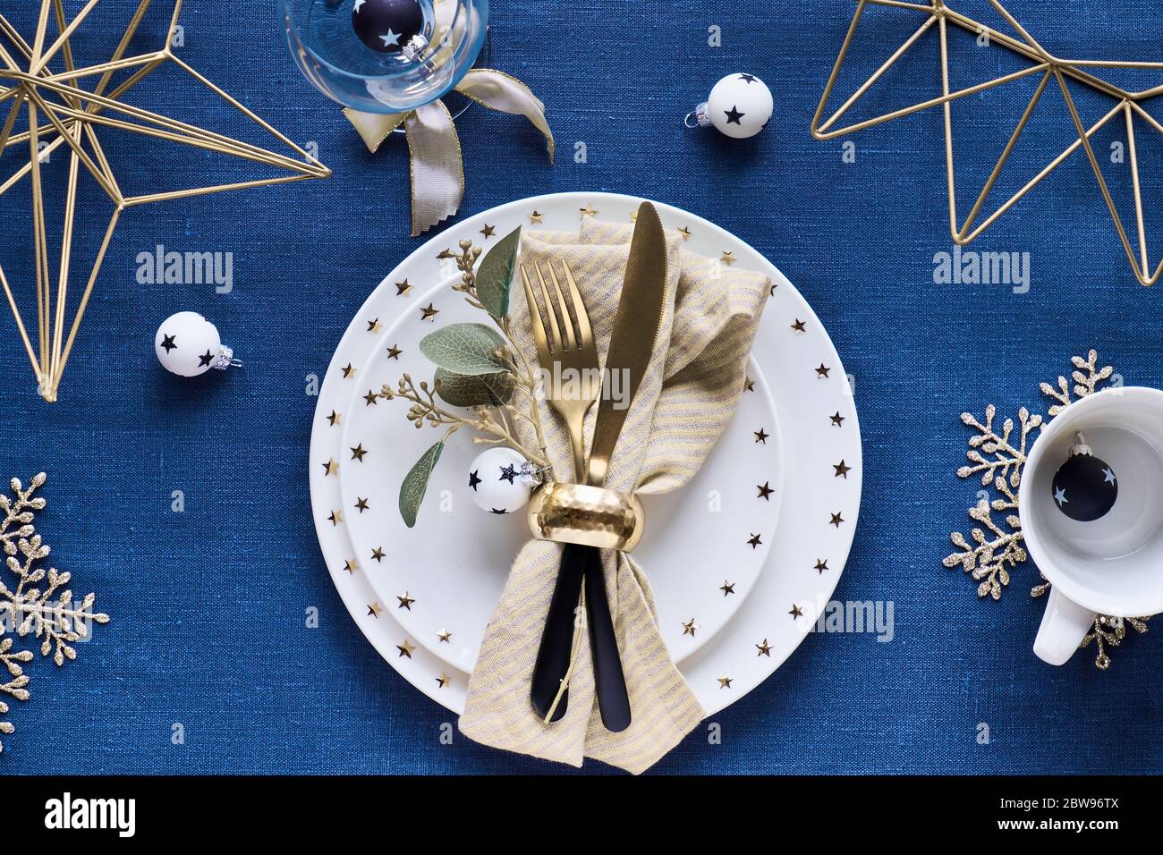 Tavolo natalizio con piatto bianco, utensili dorati e decorazioni in metallo geometrico dorato e fiocchi di neve dorati. Piatto su tessuto di lino blu scuro ta Foto Stock