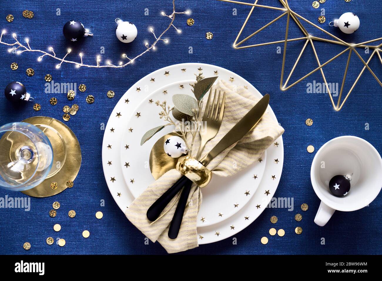 Tavolo natalizio con piatto bianco, utensili dorati e decorazioni in metallo geometrico dorato. Piatto posato su sfondo di tela di lino blu scuro di tessuto da tavolo Foto Stock