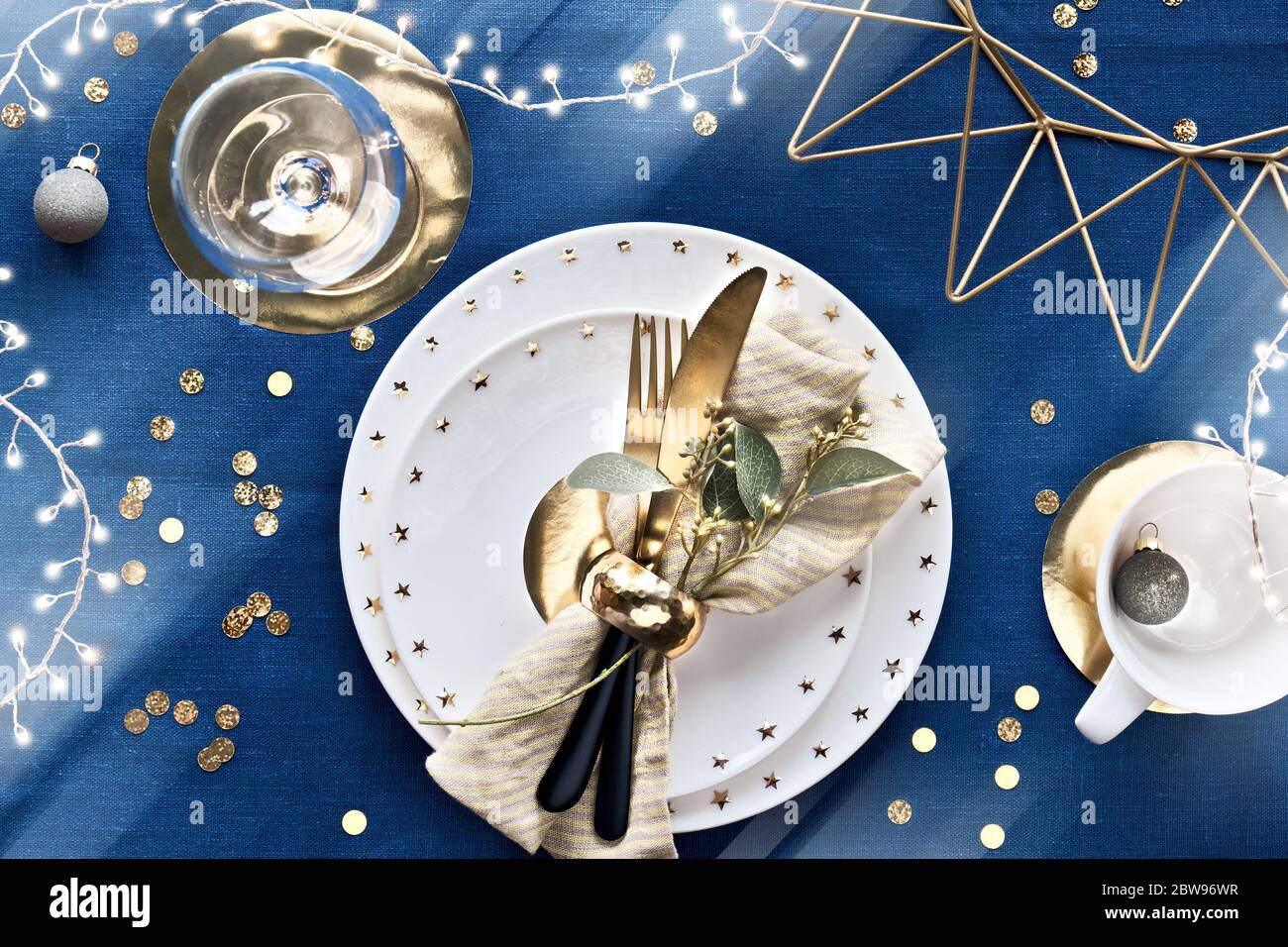 Tavolo natalizio con piatto bianco, utensili dorati e decorazioni in metallo geometrico dorato e carta. Disposizione piatta, vista dall'alto su tessuto di lino blu scuro Foto Stock