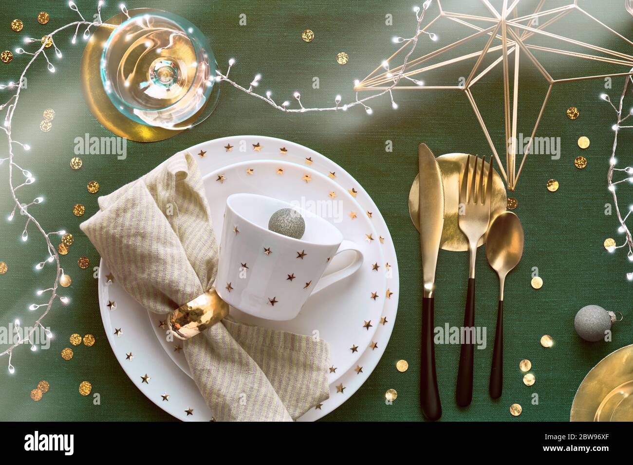 Tavolo natalizio con piatto bianco, utensili dorati e decorazioni dorate. Disposizione piatta, vista dall'alto su sfondo in tessuto di lino verde scuro. Luci natalizie Foto Stock