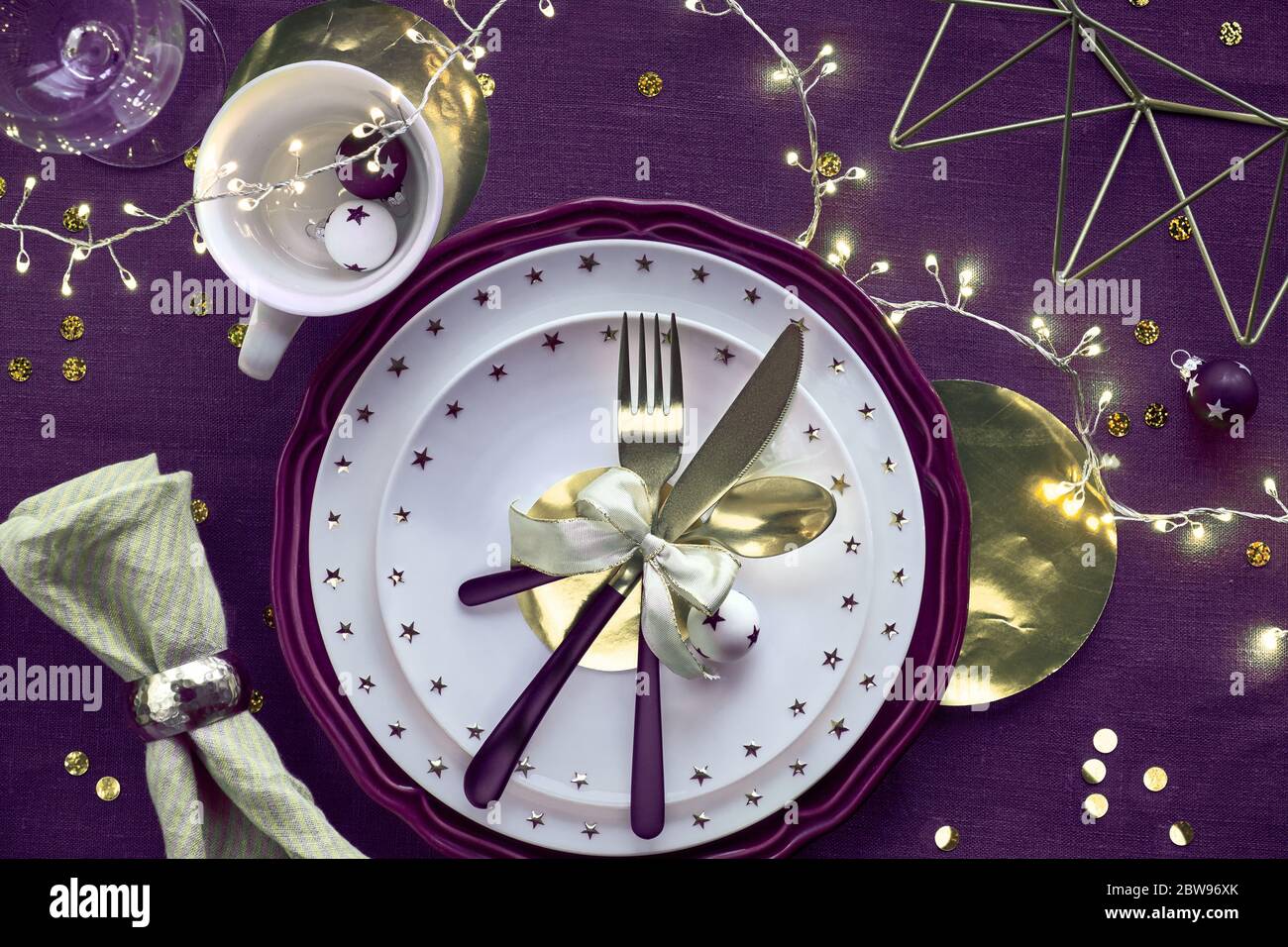 Tavolo natalizio con piatti viola e bianchi, utensili dorati e garland leggero. Disposizione piatta, vista dall'alto su fondo in tessuto di lino viola al neon scuro Foto Stock
