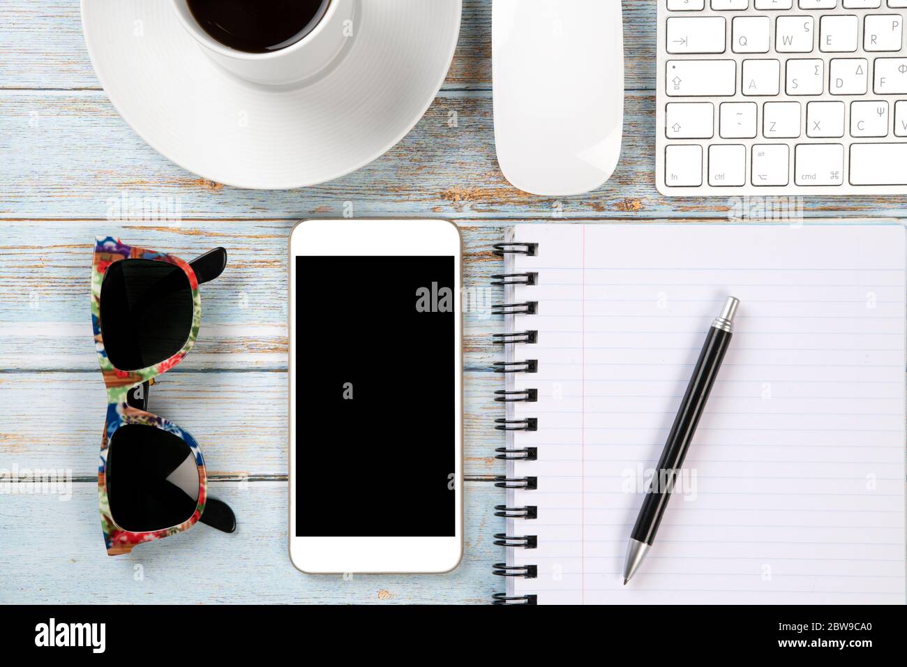 Scrivania in legno con vista dall'alto, mobile, tastiera, mouse, una tazza di caffè, notebook e occhiali da sole colorati, pronta per il lavoro Foto Stock