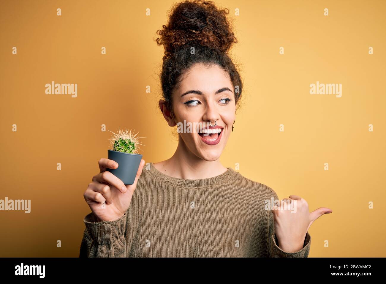 Giovane bella donna con capelli ricci e piercing tenere pentola con pianta cactus puntamento e mostrando con il pollice fino al lato con felice smilin viso Foto Stock