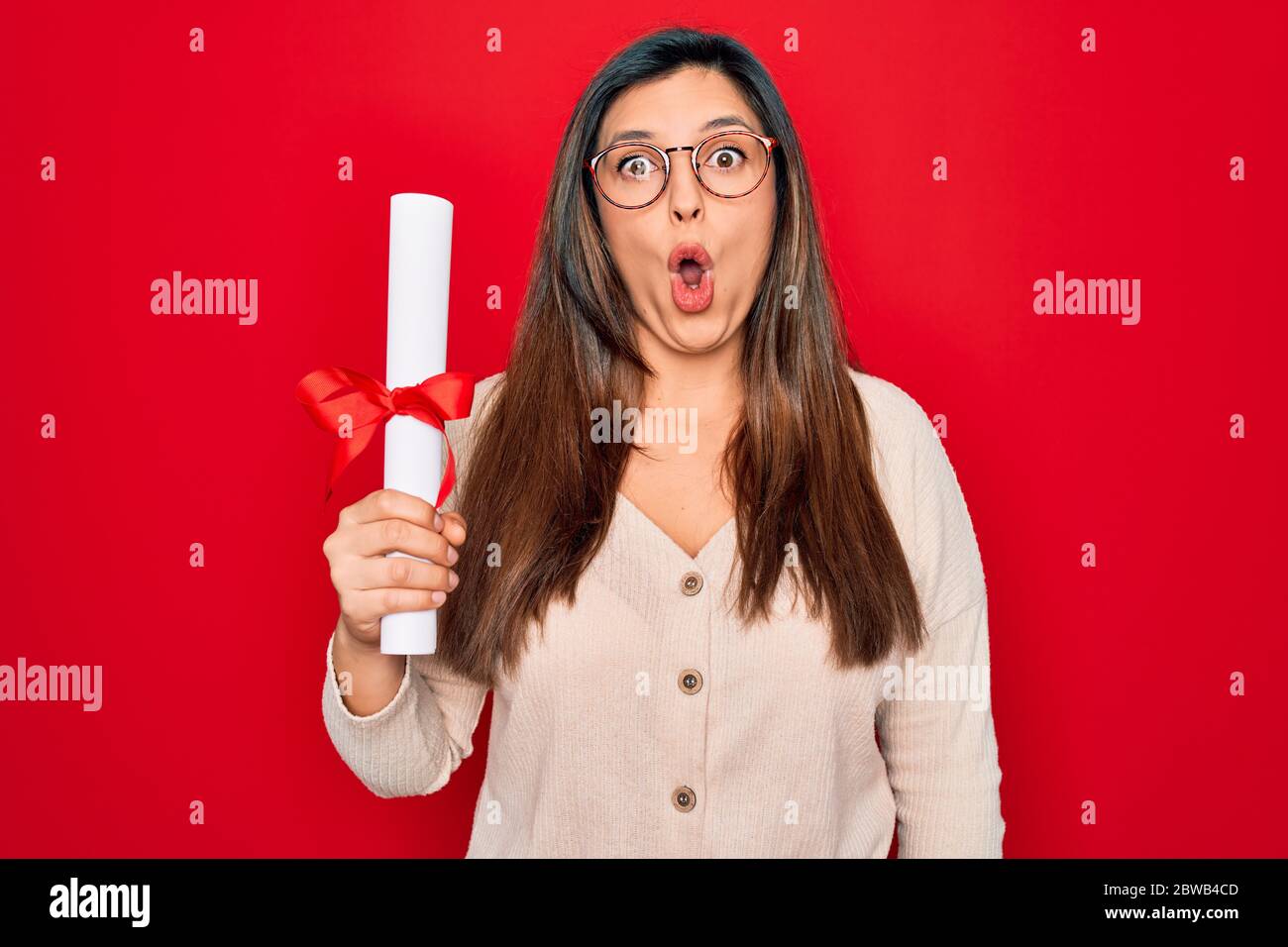 Giovane donna intelligente ispanica con occhiali che tengono la laurea universitaria su sfondo rosso spaventato in shock con un viso a sorpresa, paura ed eccitato con Foto Stock