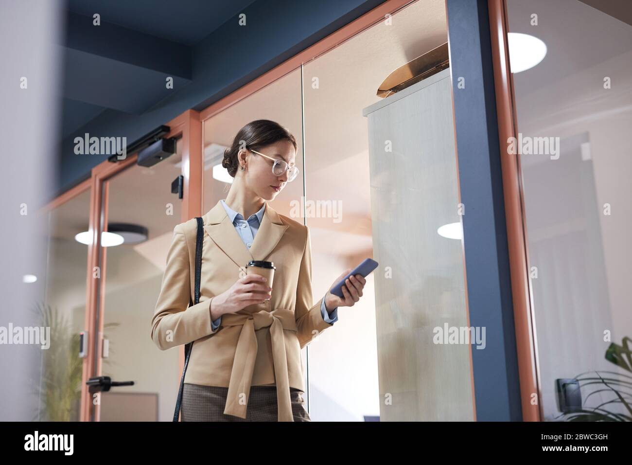 Vita ritratto di giovane donna d'affari elegante che indossa occhiali e guardando lo schermo dello smartphone mentre si cammina in contemporanea ufficio interni, copia Foto Stock