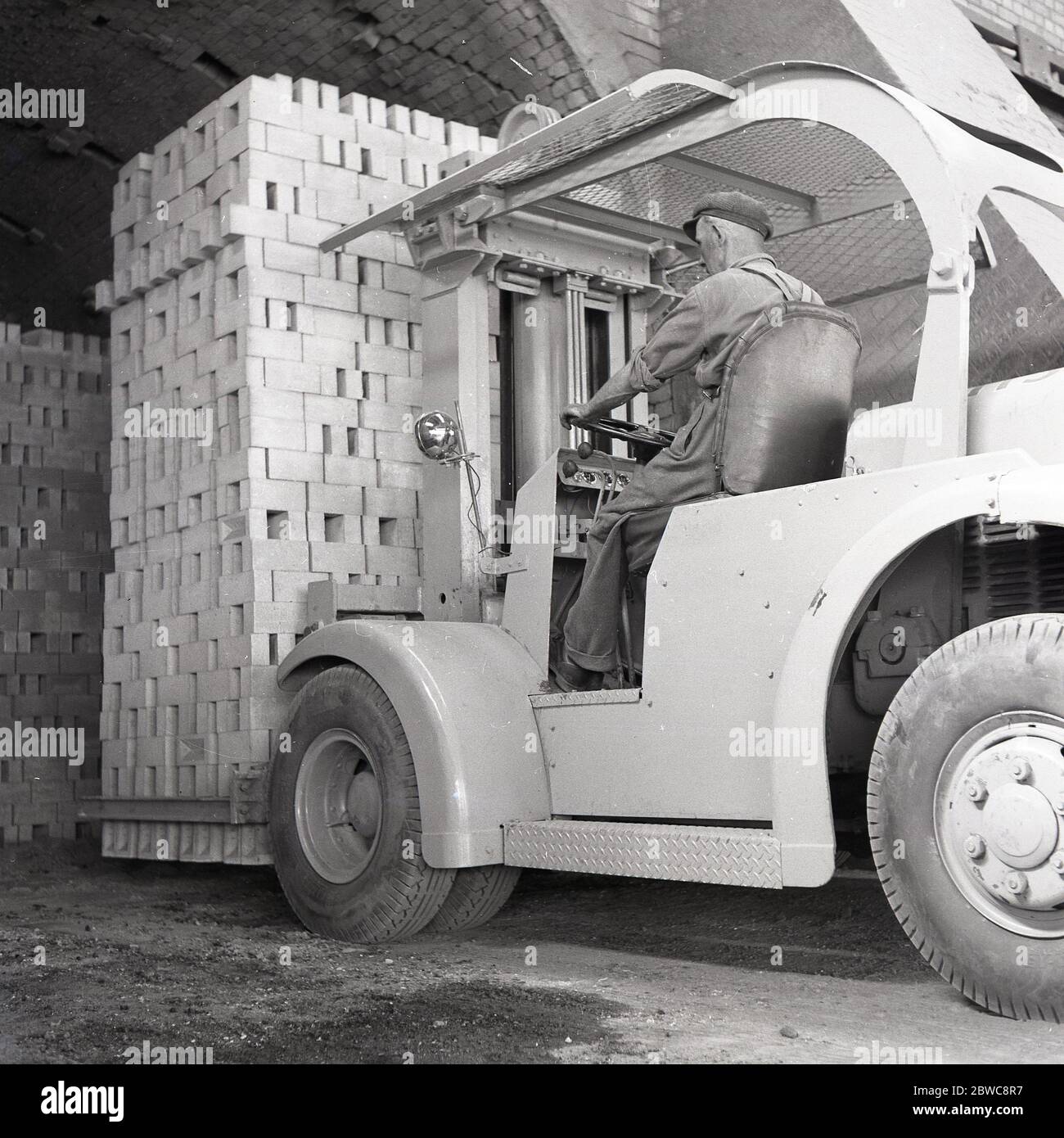 Anni '50, storico operatore maschile seduto al volante di un carrello elevatore dell'epoca che muove un carico pesante di mattoni di Londra di nuova produzione, Stewartby, Bedfordshire, Inghilterra, Regno Unito. I carrelli elevatori a batteria erano carrelli industriali a cabina aperta utilizzati per spostare materiali a breve distanza. Foto Stock