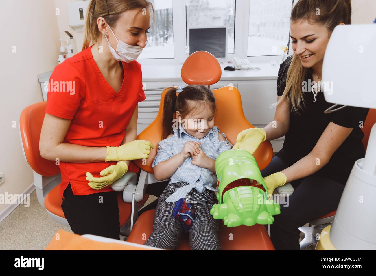 Alto angolo di felice dottore e assistente che mostra coccodrillo giocattolo a allegra bambina durante l'appuntamento in clinica contemporanea Foto Stock