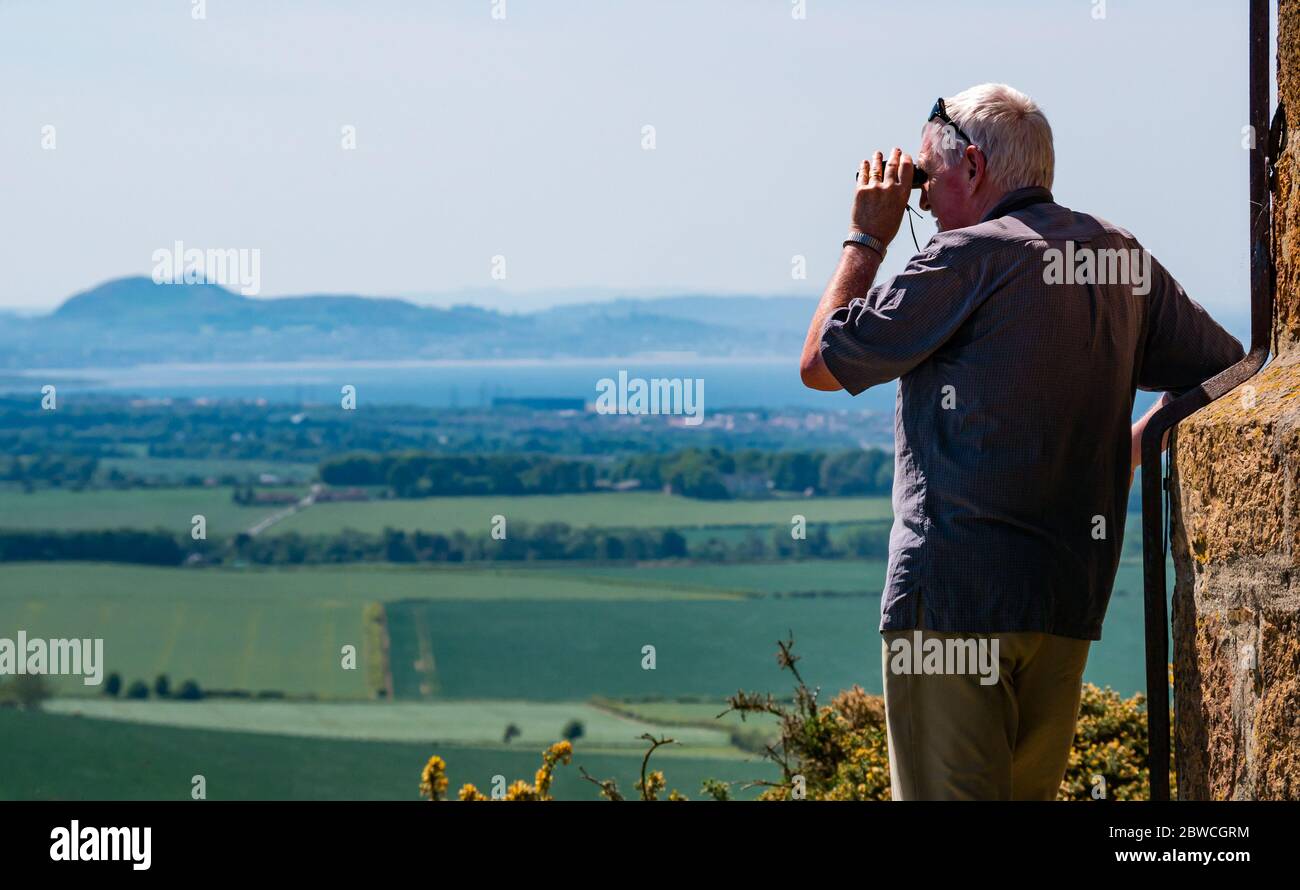 East Lothian, Scozia, Regno Unito, 31 maggio 2020. Regno Unito Meteo: Più caldo tempo in tutta la contea, ma con il sole frizzante. Un uomo anziano guarda attraverso il binocolo dalla cima di Byres Hill con il profilo distintivo di Arthur's Seat in lontananza Foto Stock