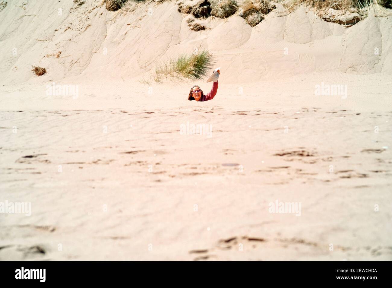 Immagine distante di donna asiatica che indossa occhiali da sole che ondolano cappello, sommerso fino al collo sotto dune di sabbia sulla spiaggia concetto: Affondare sensazione, sabbia veloce. Foto Stock