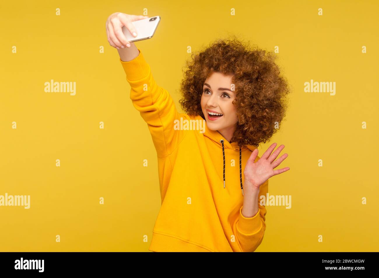 Ritratto di felice trendy ricci-capelli donna in stile urbano felpa con cappuccio prendendo selfie, avendo conversazione su videochiamata e ciao gesto ondulato, vlog broma Foto Stock