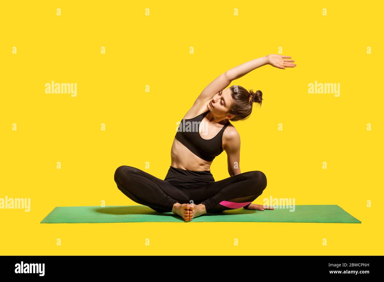 Donna sottile con capelli in pelo in intimo sportivo seduta in posizione lotus sul tappetino praticare yoga, piegandosi a fianco con la mano sollevata, facendo stretching lavoro Foto Stock