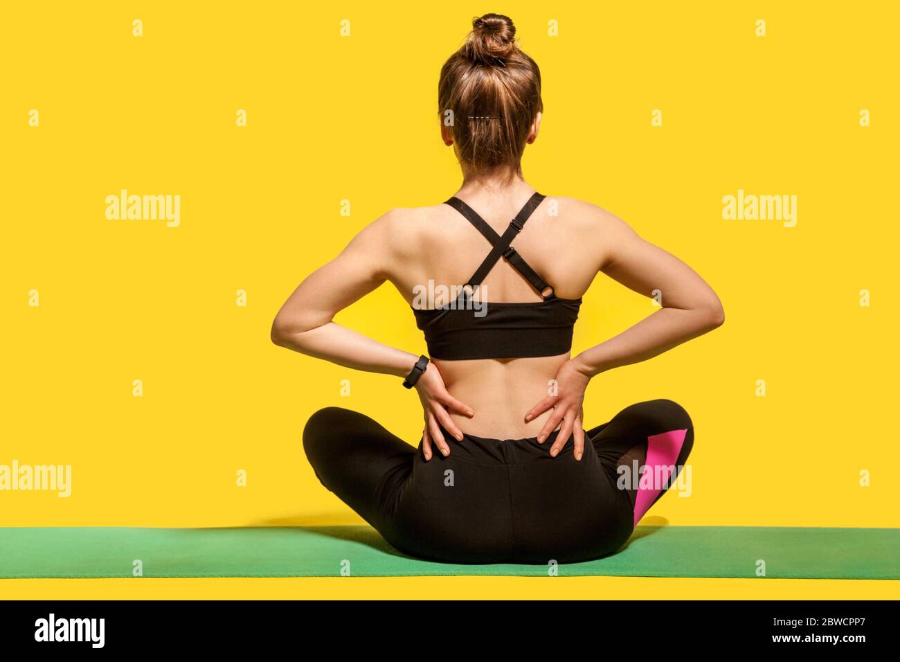 Vista posteriore, donna sportiva seduta sul tappetino e toccando la colonna vertebrale irritata, soffrendo mal di schiena dopo allenamento fisico o pratica yoga, massaggiando i muscoli rigidi Foto Stock