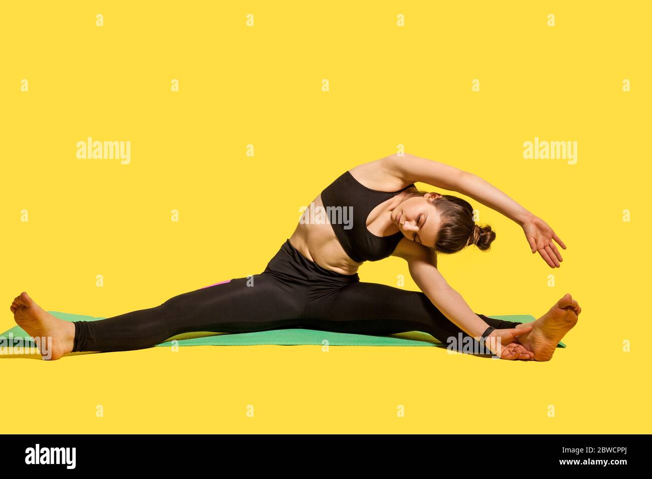Donna ginnastica sportiva con pelo in pelo sportivo seduta su tappetino con gambe spalmabili, piegandosi al dito, allungando i muscoli della schiena e delle mani, flexi Foto Stock