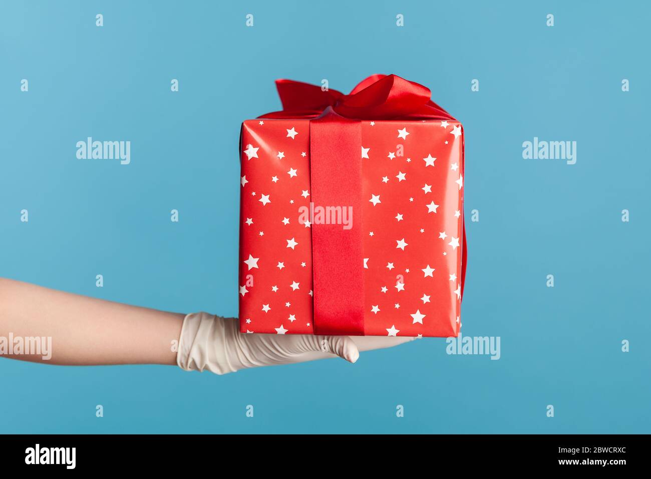 Profilo Vista laterale primo piano della mano umana in guanti chirurgici bianchi che tengono una confezione regalo rossa. Concetto di condivisione, donazione o consegna. Interno, studio shot, isola Foto Stock