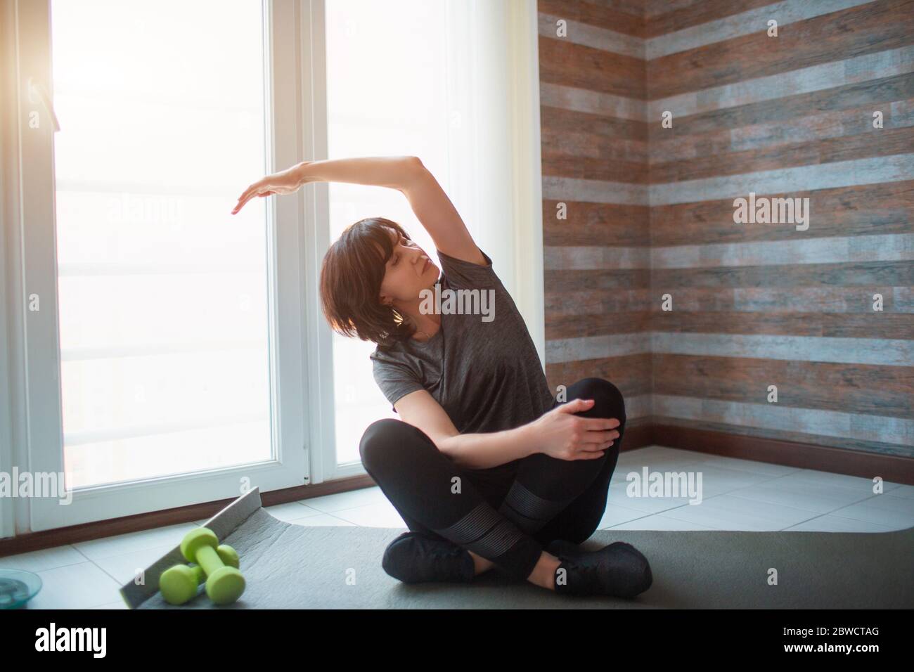 Donna slim fit adulta si allena a casa. Modello senior con gambe incrociate e allunga il braccio a lato. Esercitandosi da solo per ottenere in buona forma e. Foto Stock