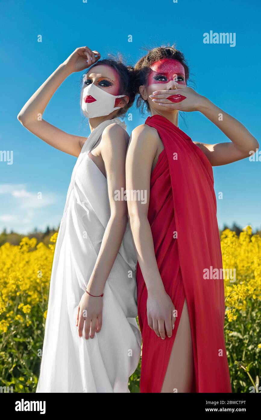 Due belle ragazze di brunette con trucco creativo luminoso in tunics su uno sfondo di un campo di fiori gialli e cielo blu. Una ragazza in una maschera Foto Stock