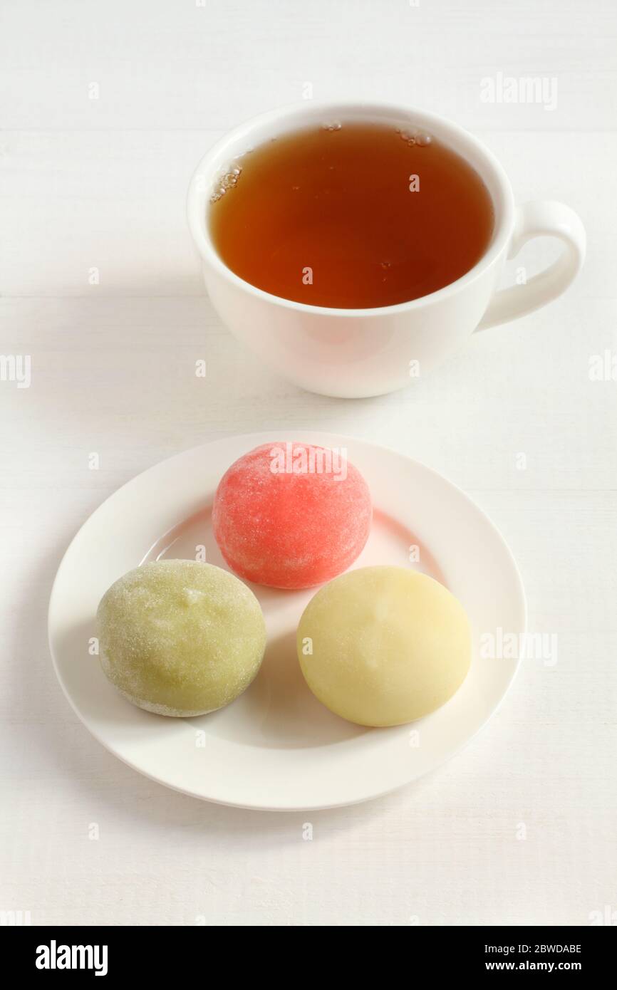 Tre tipi di dessert giapponesi mochi - melograno al miele, tè verde matcha, frutto della passione su un piatto bianco e una tazza con tè su un legno bianco Foto Stock