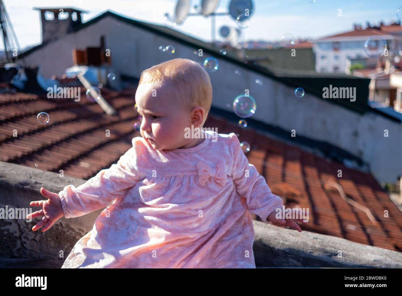 Primo piano di cute baby girl indossare adorabile vestito sul tetto, bolle di sapone galleggianti intorno, bambina cercando di prendere le bolle Foto Stock