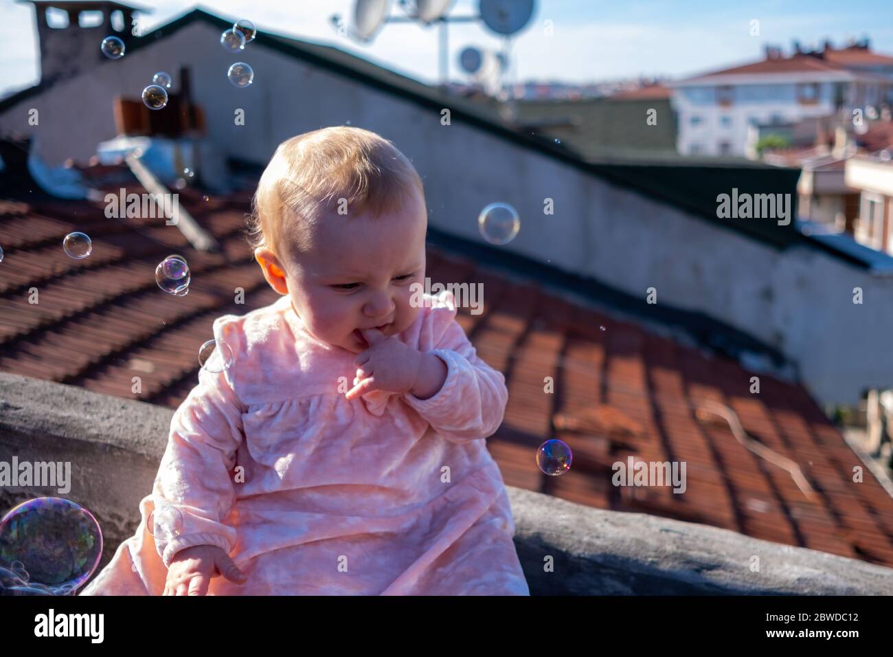Primo piano di cute baby girl indossare adorabile vestito sul tetto, bolle di sapone galleggianti intorno, bambina cercando di prendere le bolle Foto Stock