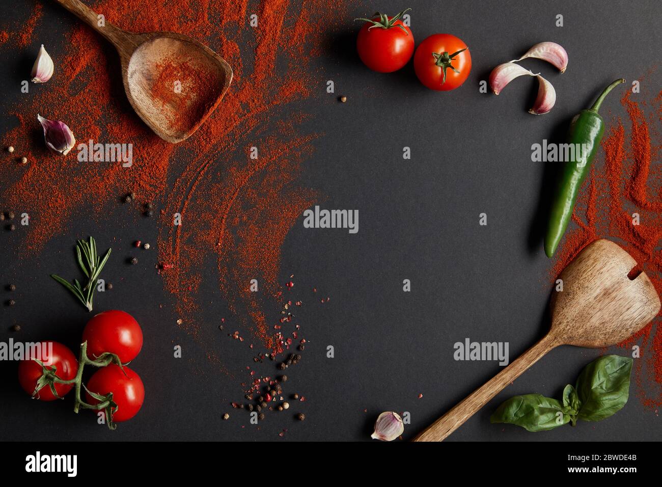 vista dall'alto di pomodori ciliegini rossi, peperoncino piccante, spicchi d'aglio ed erbe fresche vicino a cucchiai con paprika in polvere su nero Foto Stock